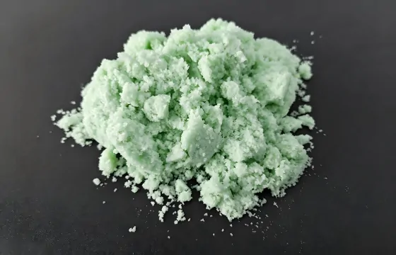 该硫酸亚铁常温下为浅蓝绿色结晶粉末，结块成堆，夹杂细碎颗粒，周边散落细料，无明显特殊气味。