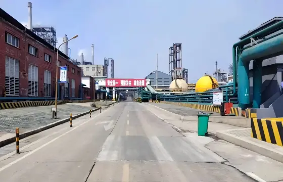 化工厂区主干道，两侧分布红砖墙厂房、大型管道与球形储罐，道路上方悬挂安全生产红色标语横幅，纯碱主要应用于玻璃、洗涤剂、冶金、造纸、印染、食品、医药及水处理等行业。